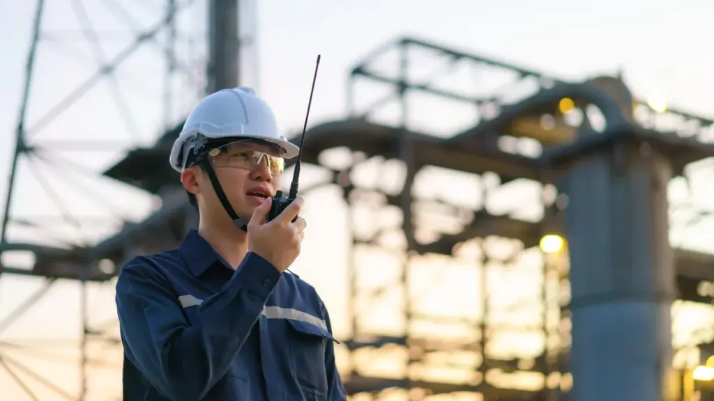 Técnico de capacete fala por rádio diante das estruturas metálicas de uma refinaria ao entardecer